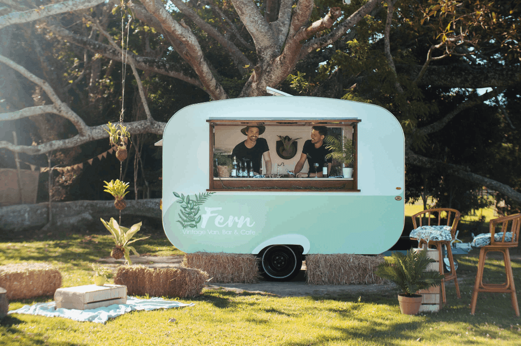 Two bartenders inside the mobile bar of Fern Vintage Van serving a woman with drinks.