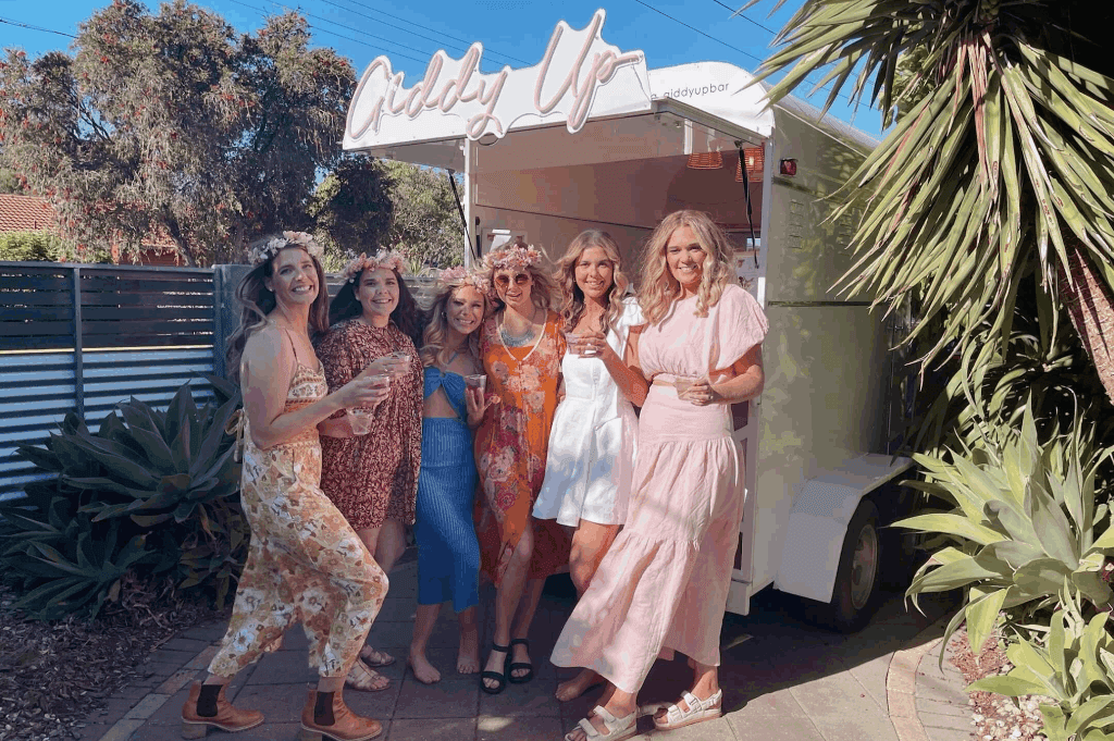 Six women wearing floral dresses posing infront of the mobile bar holding drinks.